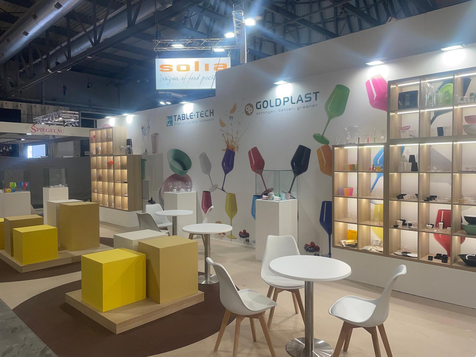Stand GOLDPLAST con tavoli circolari, cubi gialli e scaffalature in legno